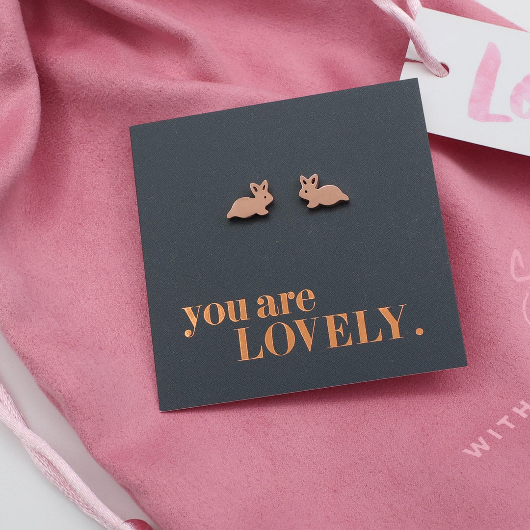 Rose gold bunny earrings on a dark card with 'you are LOVELY.' text, against a pink fabric background.