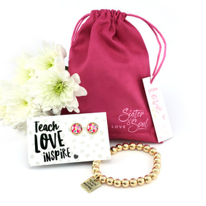 A hot pink velour gift bag with 'TEACH LOVE INSPIRE' printed on a card, alongside a gold hematite bracelet and gold circle stud earrings, presented with decorative white flowers.