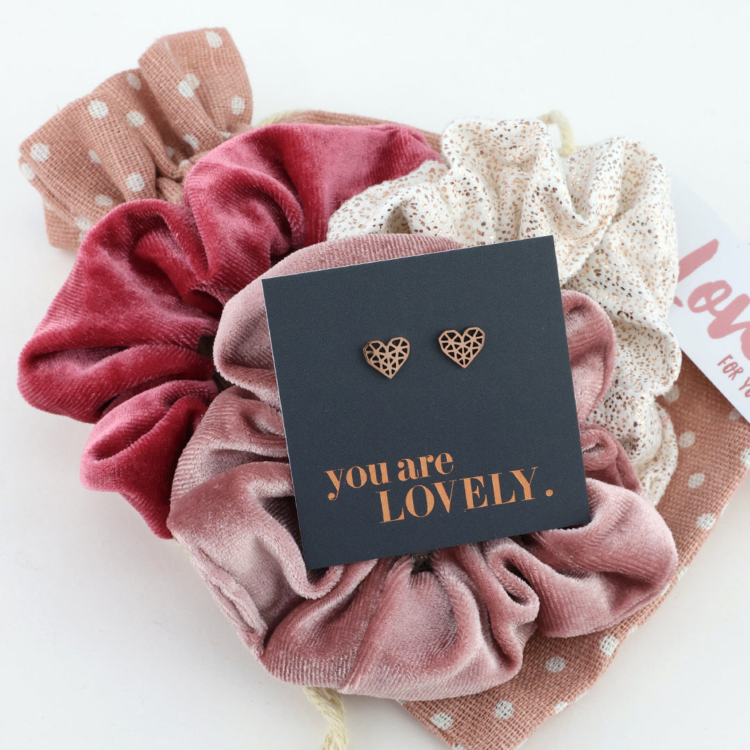 Pink scrunchies with heart-shaped earrings and a 'you are LOVELY.' card on a white background