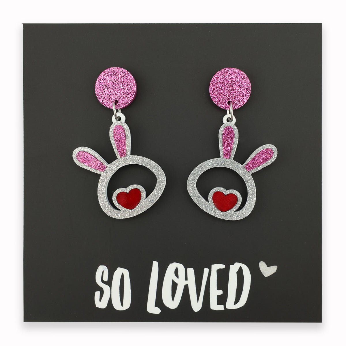 A pair of acrylic statement dangle earrings in the shape of bunny faces with pink glittery tops and red hearts, displayed on a black background with the text 'SO LOVED'.