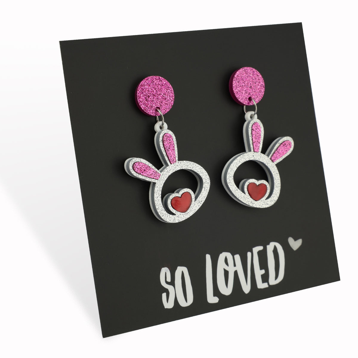 A pair of acrylic statement dangle earrings in the shape of bunny faces with pink glittery tops and red hearts, displayed on a black background with the text 'SO LOVED'.