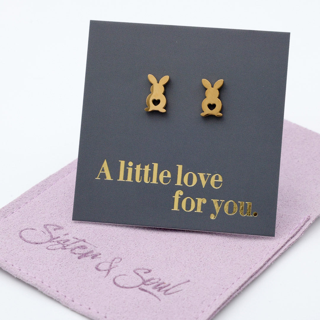 Gold bunny-shaped earrings on a dark gray card with 'A little love for you' text, placed on a pink surface with 'Sister & Soul' branding.