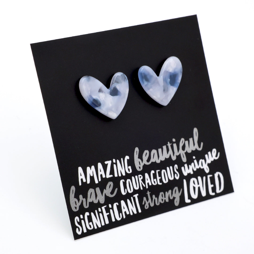 A pair of heart-shaped stud earrings with a cloudy, marbled light blue and white appearance, displayed on a black square card with inspirational text.