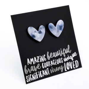 A pair of heart-shaped stud earrings with a cloudy, marbled light blue and white appearance, displayed on a black square card with inspirational text.