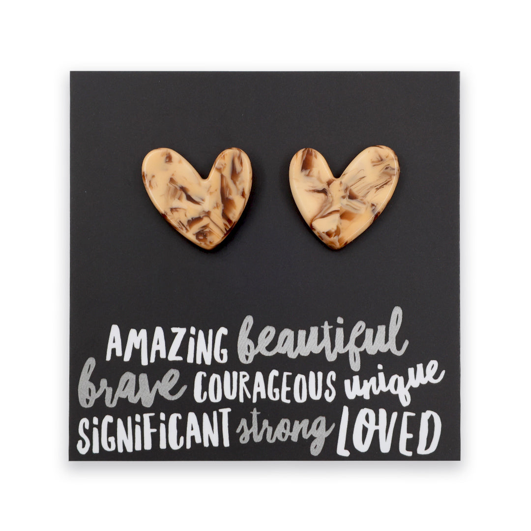 A pair of heart-shaped stud earrings with a marbled beige and brown wood-grain pattern, displayed on a black square card with white inspirational text.