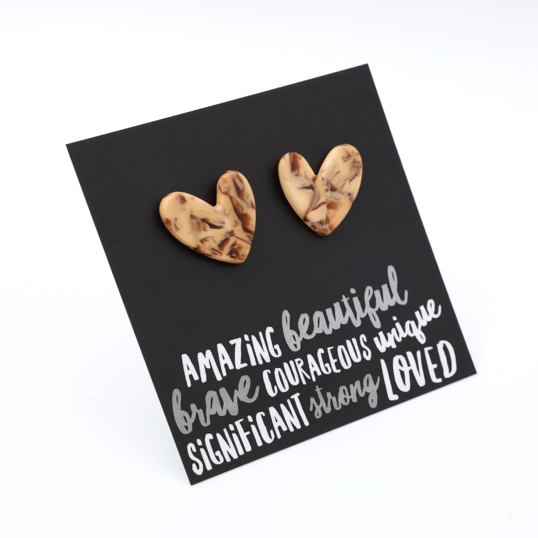 A pair of heart-shaped stud earrings with a marbled beige and brown wood-grain pattern, displayed on a black square card with white inspirational text.