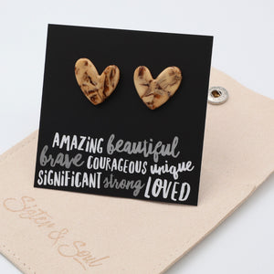 A pair of heart-shaped stud earrings with a marbled wood pattern, displayed on a black card with inspirational text, resting on a beige pouch.