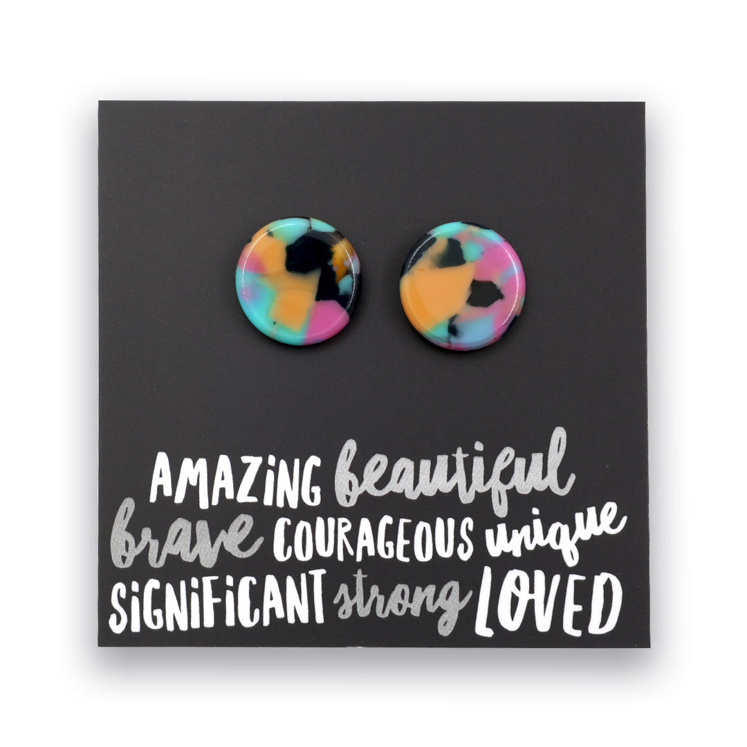A pair of round stud earrings with a colorful marbled pattern in pink, yellow, blue, and black, displayed on a black card with white text that reads 'AMAZING beautiful brave COURAGEOUS unique SIGNIFICANT strong LOVED'.