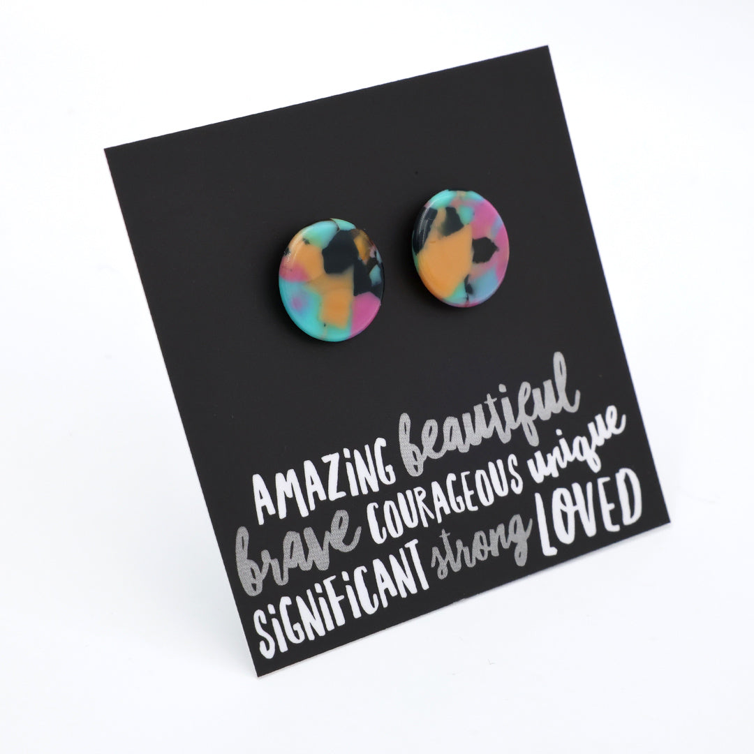 A pair of round stud earrings with a colorful marbled pattern in pink, yellow, blue, and black, displayed on a black card with white text that reads 'AMAZING beautiful brave COURAGEOUS unique SIGNIFICANT strong LOVED'.