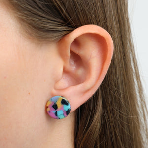A close-up of a person's ear wearing a round stud earring with a colorful marbled pattern in pink, blue, yellow, black, and white.