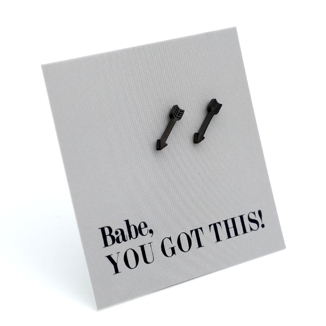 A pair of black arrow-shaped stud earrings displayed on a textured light gray square card that reads 'Babe, YOU GOT THIS!' in black text, photographed on a white background.