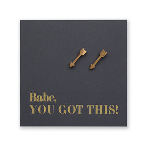 A pair of small gold arrow-shaped stud earrings displayed on a dark charcoal square backing card with the phrase 'Babe, YOU GOT THIS!' in gold lettering, shown against a plain white background.