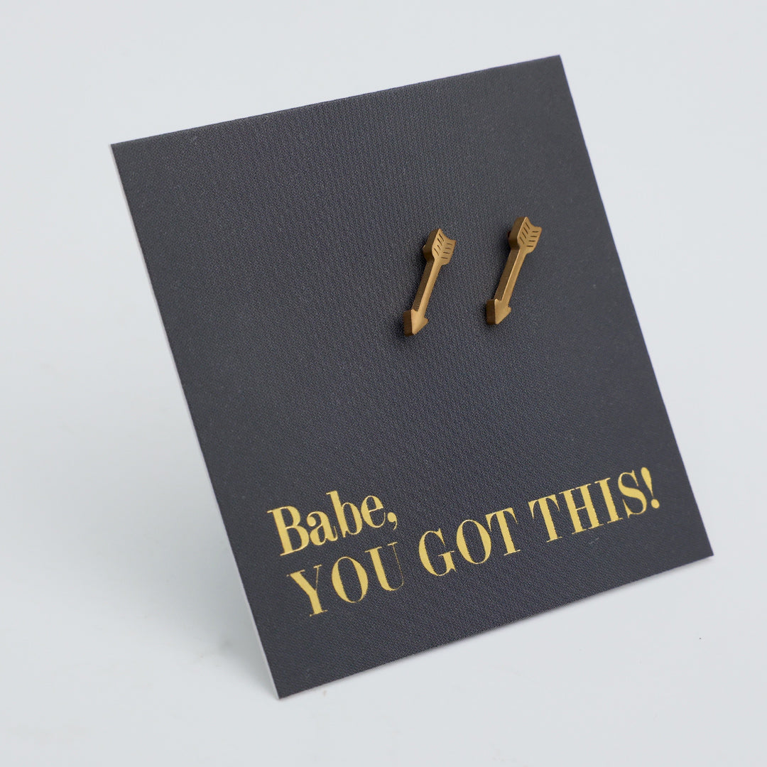 A pair of small gold arrow-shaped stud earrings displayed on a dark charcoal textured backing card with gold text that reads 'Babe, YOU GOT THIS!' on a light gray background.