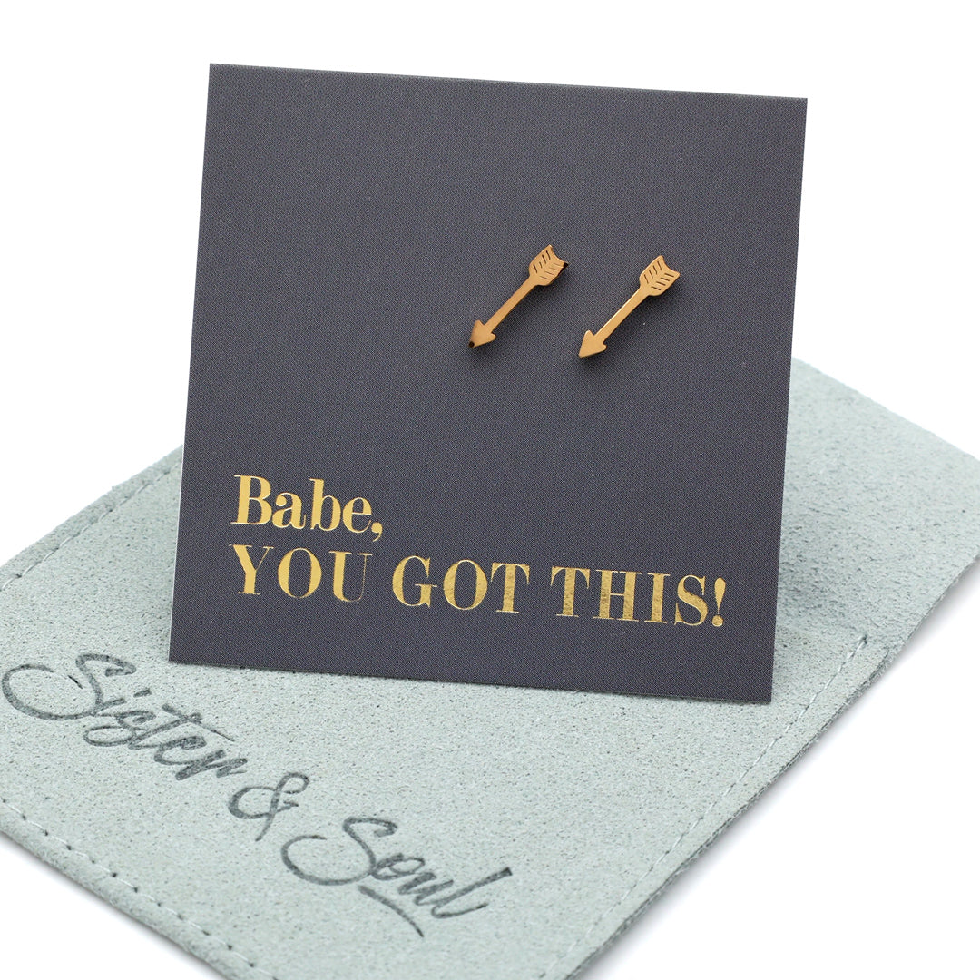 Two gold arrow-shaped stud earrings displayed on a dark charcoal card that reads 'Babe, YOU GOT THIS!' in gold lettering, resting on a light sage green jewelry pouch with 'Sister & Soul' written on it.