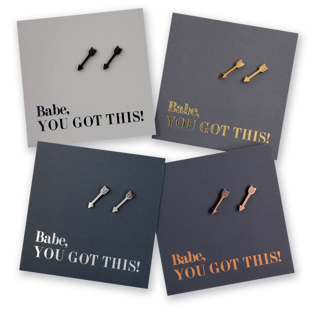 Four square product cards arranged in a 2x2 grid, each showing a pair of small arrow-shaped stud earrings on a dark card with the phrase 'Babe, YOU GOT THIS!' in metallic lettering. The earrings and cards come in black, gold, silver, and rose gold colorways.
