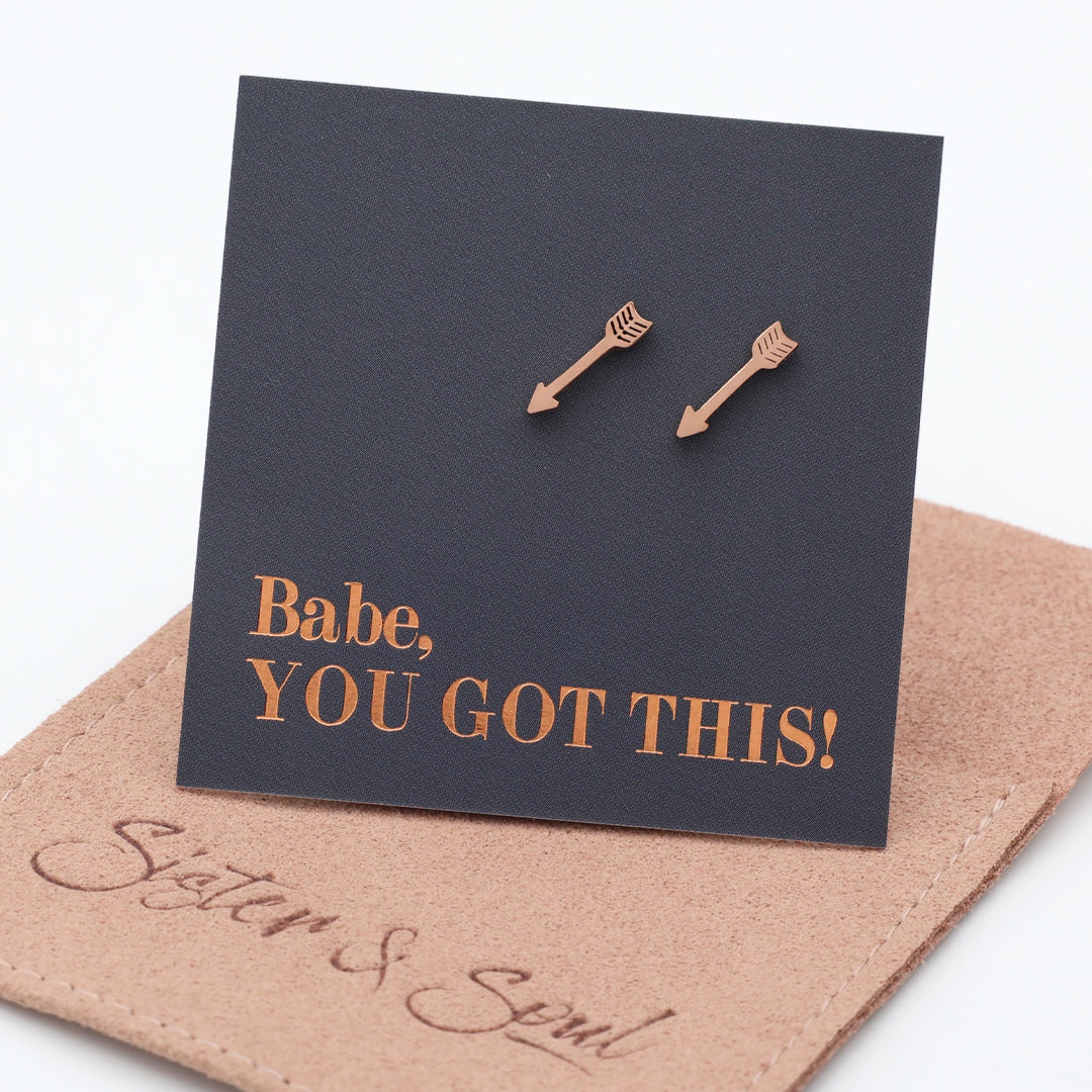 A pair of rose gold arrow stud earrings displayed on a black square backing card that reads 'Babe, YOU GOT THIS!' in rose gold lettering. The card is propped up on a  brown fabric pouch with the brand name 'Sister & Soul' embossed on it, set against a clean white background.