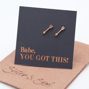 A pair of rose gold arrow stud earrings displayed on a black square backing card that reads 'Babe, YOU GOT THIS!' in rose gold lettering. The card is propped up on a  brown fabric pouch with the brand name 'Sister & Soul' embossed on it, set against a clean white background.