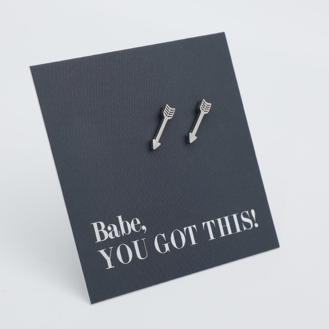 A pair of silver arrow-shaped stud earrings displayed on a dark charcoal card that reads 'Babe, YOU GOT THIS!' in white text, set against a clean light gray background.