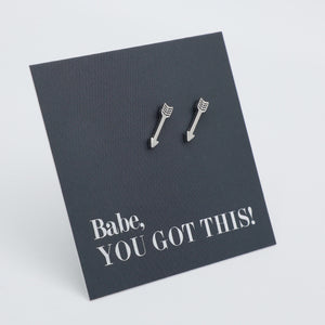 A pair of silver arrow-shaped stud earrings displayed on a dark charcoal card that reads 'Babe, YOU GOT THIS!' in white text, set against a clean light gray background.
