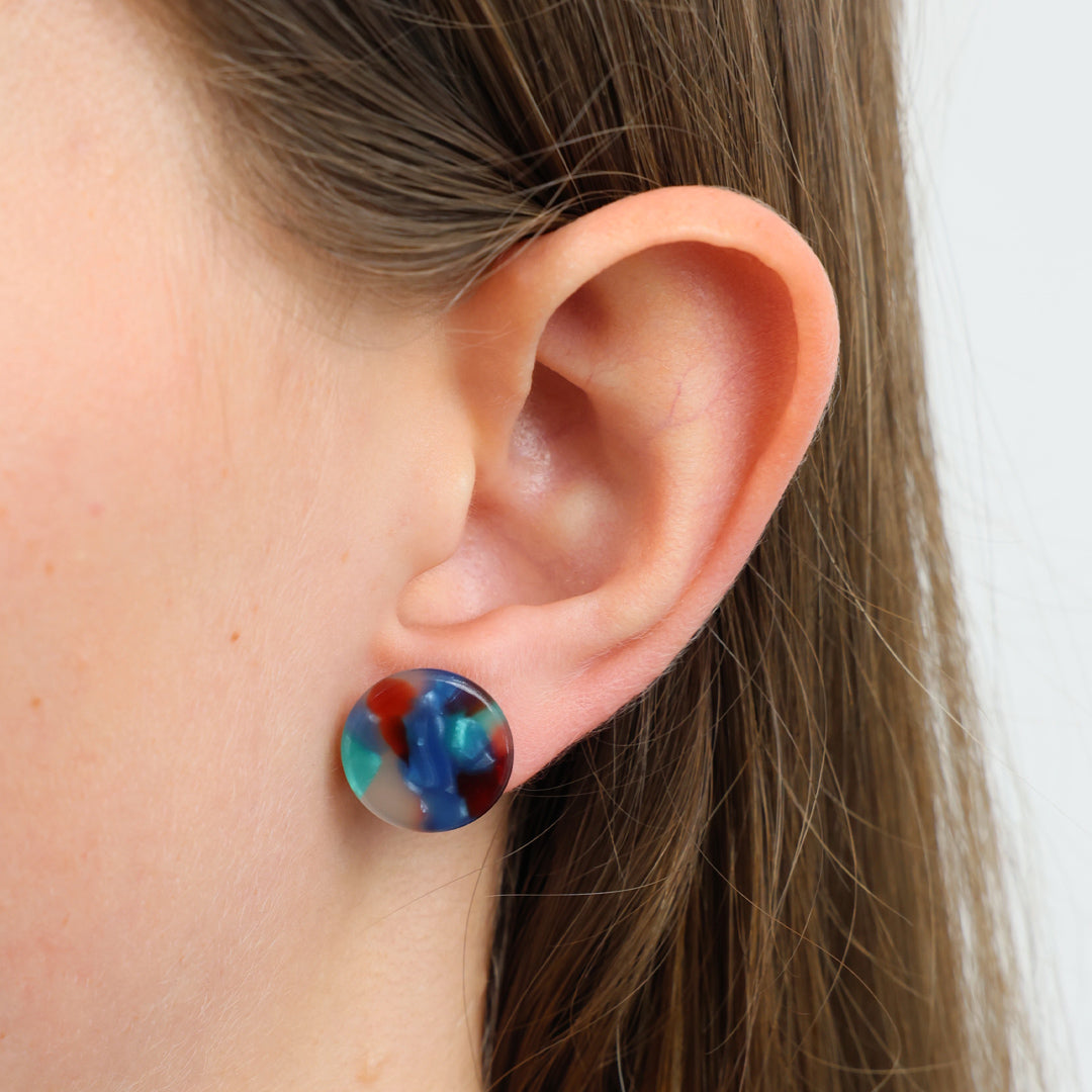 A close-up of a person's ear wearing a round stud earring with a marbled, swirled pattern of blue, red, and teal.