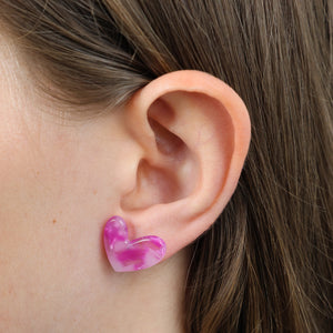 A close-up of a person's ear wearing a small heart-shaped stud earring made of marbled pink and purple resin.