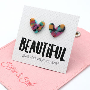 A pair of heart-shaped stud earrings with a colorful tie-dye pattern in pink, yellow, blue, and green, displayed on a white card that says 'BEAUTIFUL just the way you are!' resting on a pink fabric pouch with 'Sister & Soul' written on it.