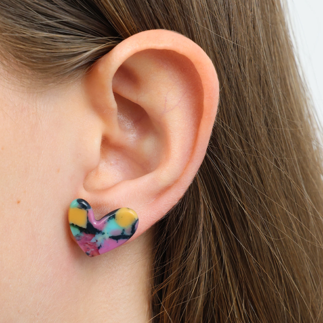 A close-up of a person's ear wearing a small heart-shaped stud earring with a colorful tie-dye pattern in pink, yellow, black, and blue.