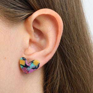 A close-up of a person's ear wearing a small heart-shaped stud earring with a colorful tie-dye pattern in pink, yellow, black, and blue.