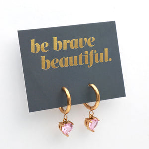 Gold huggie hoop earrings with dangling pink heart-shaped crystals, displayed on a dark gray card with gold foil text reading 'be brave beautiful.' against a white background.