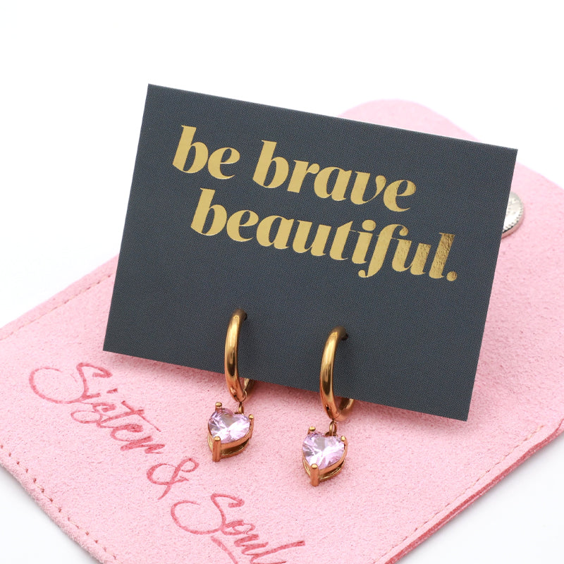 A pair of gold hoop earrings with heart-shaped pink gemstones, displayed on a black card that reads 'be brave beautiful.' resting on a pink 'System & Soul' pouch.