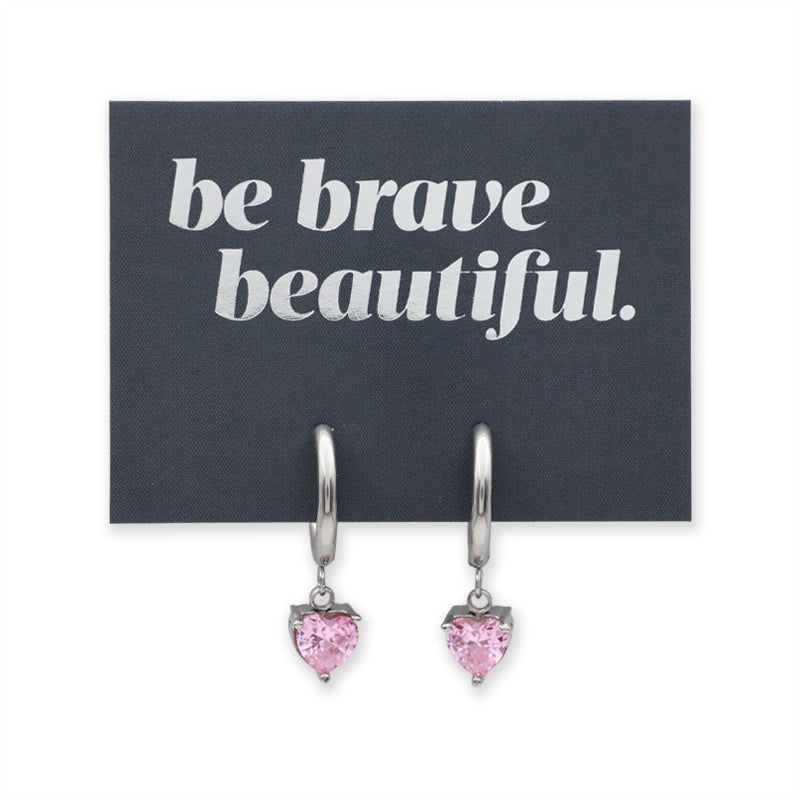 A pair of silver-toned drop earrings with small heart-shaped pink gemstones, displayed on a dark charcoal card that reads 'be brave beautiful.' in white text, against a plain white background.