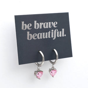 Silver huggie hoop earrings with dangling pink heart-shaped crystals, displayed on a black card that reads 'be brave beautiful.' against a white background.