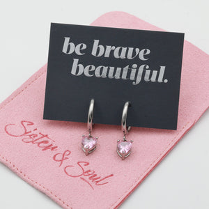 A pair of silver huggie hoop earrings with heart-shaped pink gemstones, displayed on a black card that reads 'be brave beautiful.' resting on a pink velvet jewelry pouch branded 'Sister & Soul'.