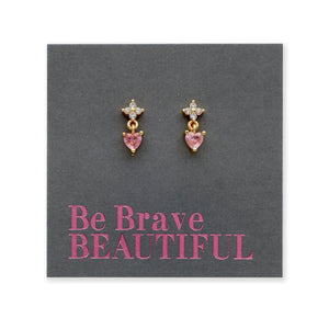 A pair of gold dangle earrings with small clear stones at the top and pink heart-shaped stones below, displayed on a dark gray backing card that reads 'Be Brave BEAUTIFUL' in pink text.