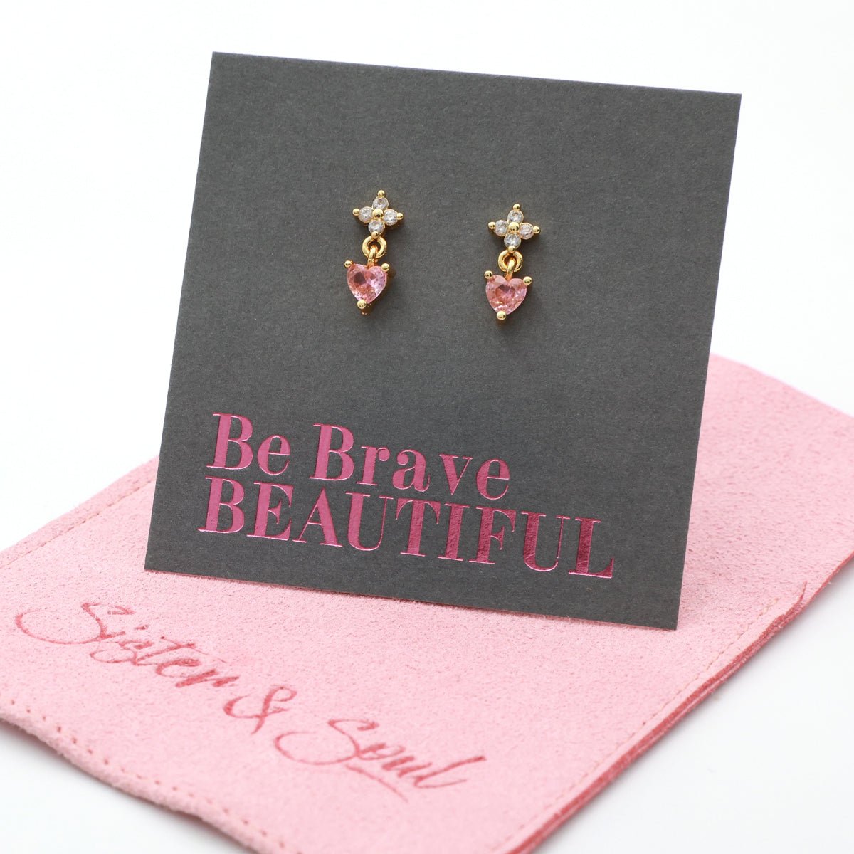 Gold-tone dangle earrings with a small clear crystal star and a pink heart-shaped crystal drop, displayed on a dark gray card that reads 'Be Brave BEAUTIFUL' above a pink branded jewelry pouch.