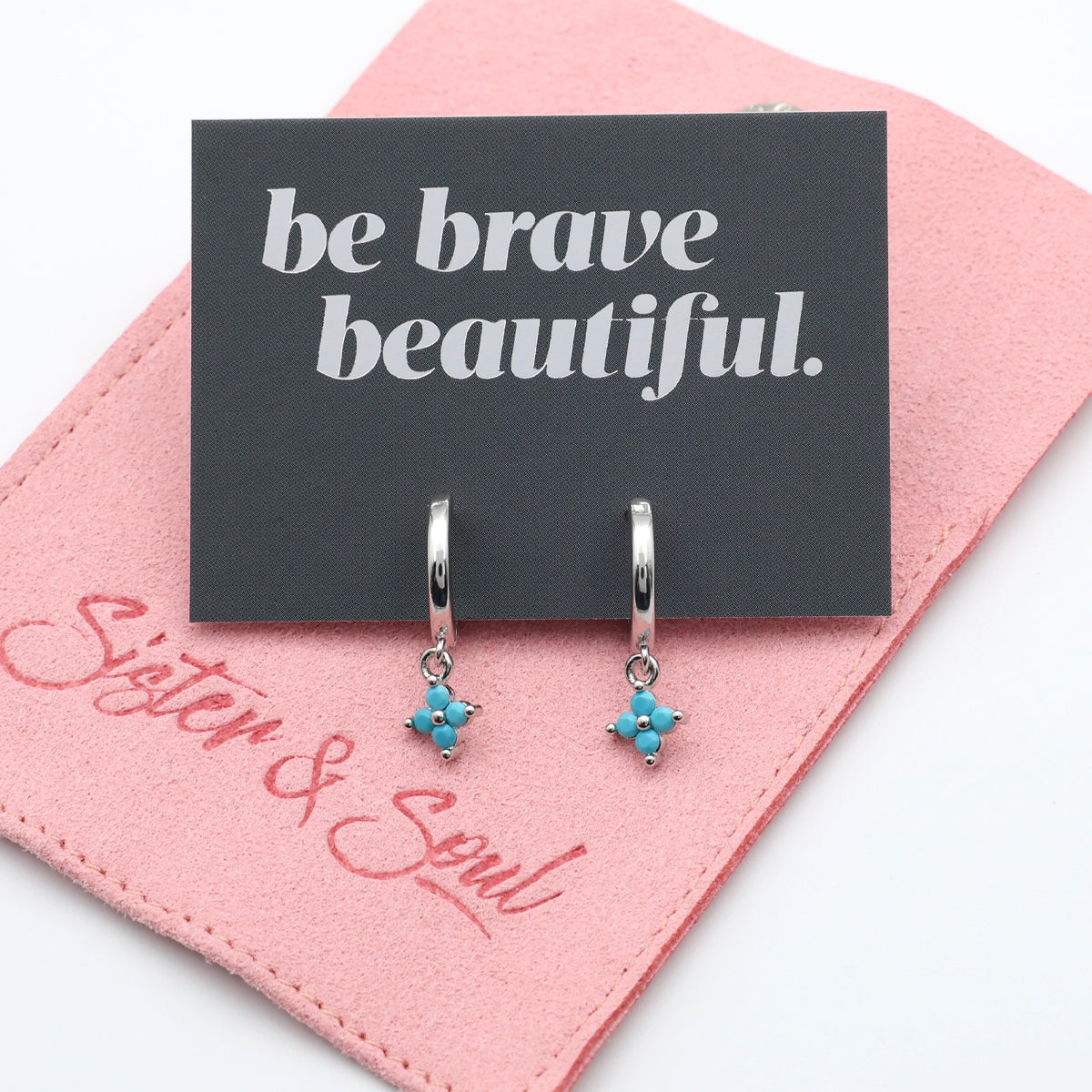 Silver earrings with blue accents on a 'be brave beautiful' card with pink background