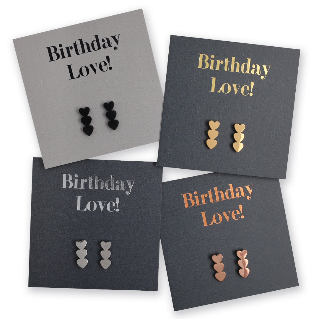 Four square jewelry display cards arranged on a white background, each holding a pair of small heart-shaped stud earrings. The cards are gray, charcoal, and dark gray, with the text 'Birthday Love!' in black, gold, silver, and rose gold tones.