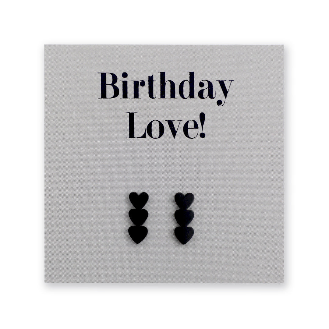 A pair of black heart-shaped stud earrings displayed on a dark charcoal gray gift card that reads 'Birthday Love!' in black lettering.