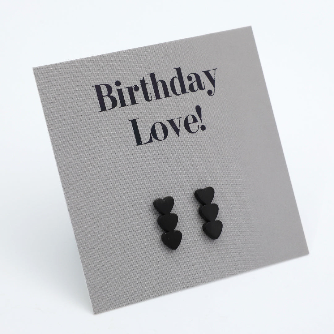 Black heart stud earrings displayed on a textured gray card that reads 'Birthday Love!' in black serif text, photographed on a clean white background.