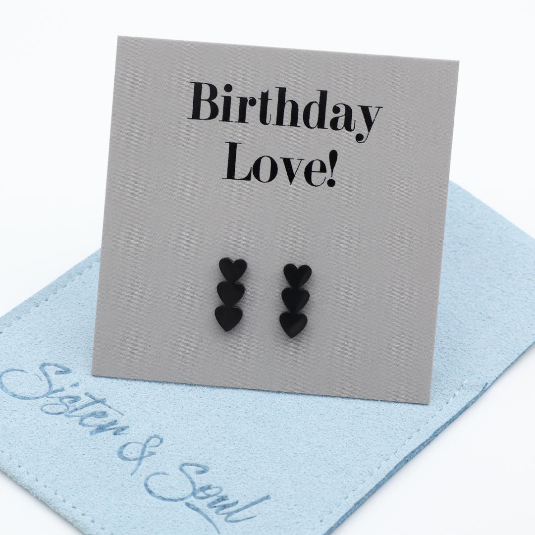 Three pairs of small black heart stud earrings displayed on a gray card that reads 'Birthday Love!' and resting on a light blue fabric pouch with 'Sister & Soul' embroidered in blue script.