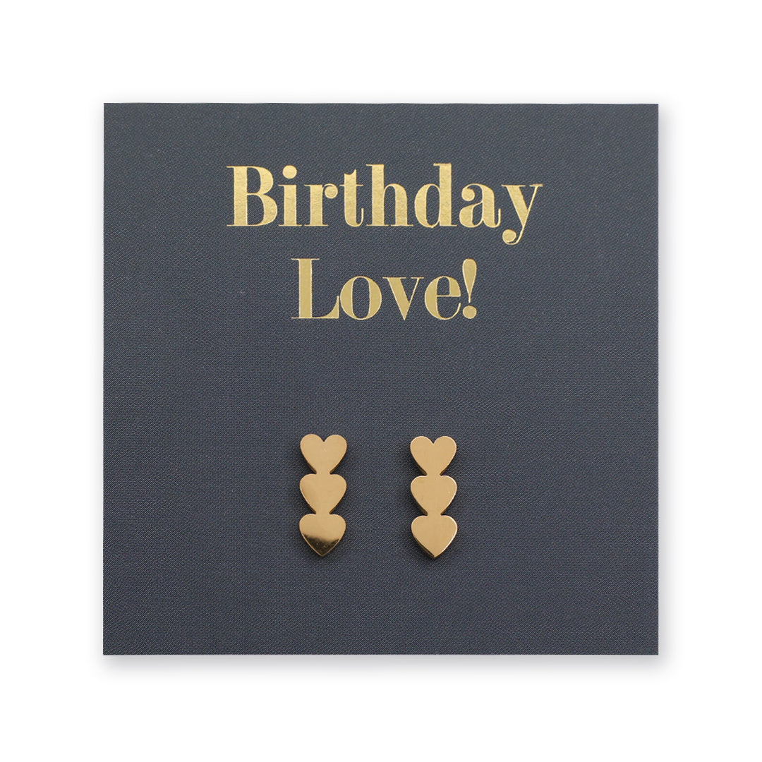 A pair of gold heart-shaped stud earrings displayed on a dark charcoal gray gift card that reads 'Birthday Love!' in gold lettering.