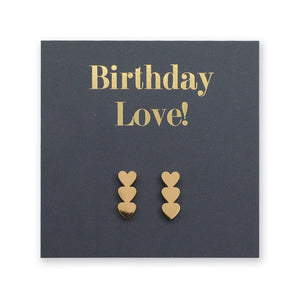 A pair of gold heart-shaped stud earrings displayed on a dark charcoal gray gift card that reads 'Birthday Love!' in gold lettering.