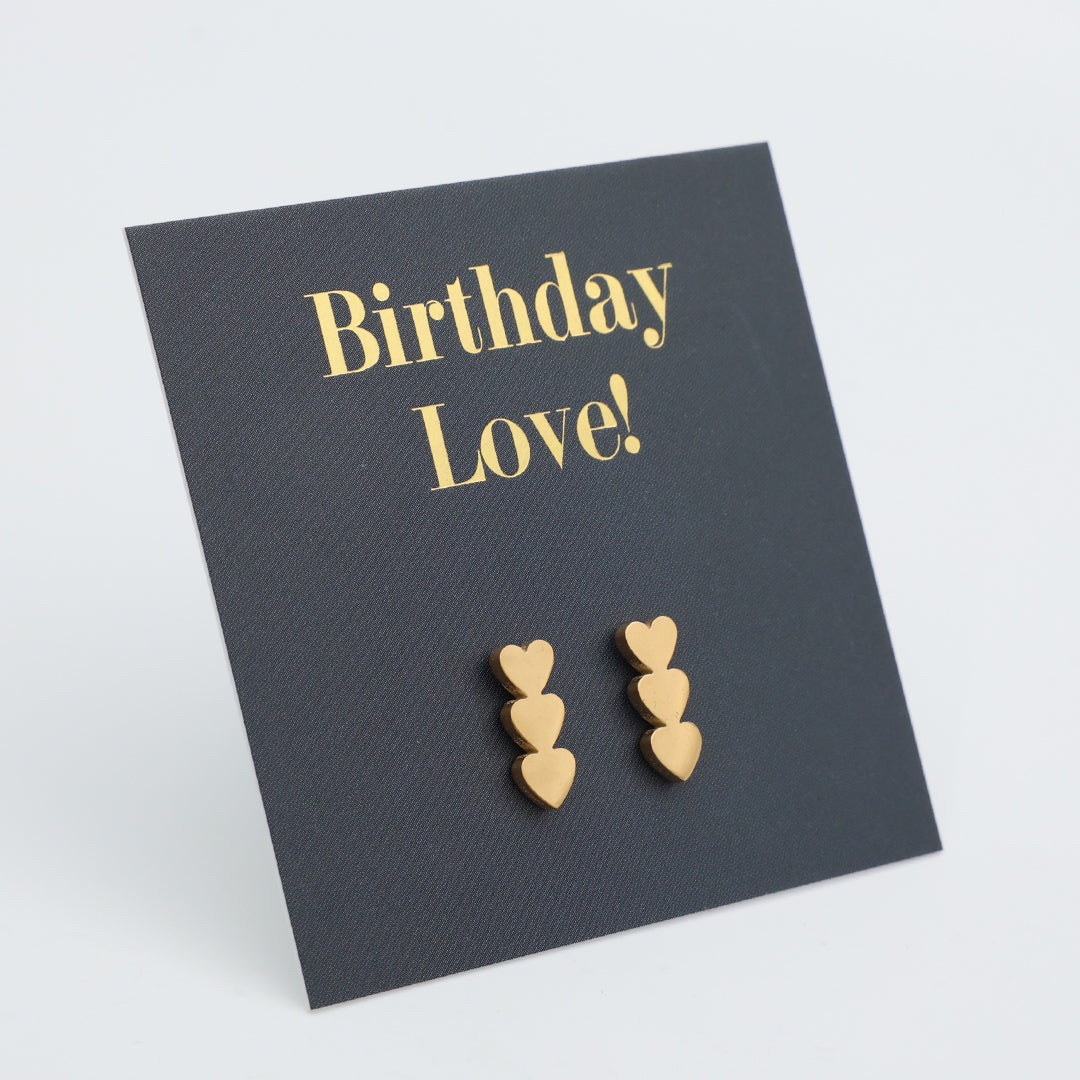 A pair of small gold heart stud earrings displayed on a dark charcoal card that reads 'Birthday Love!' in gold lettering, photographed on a light gray background.