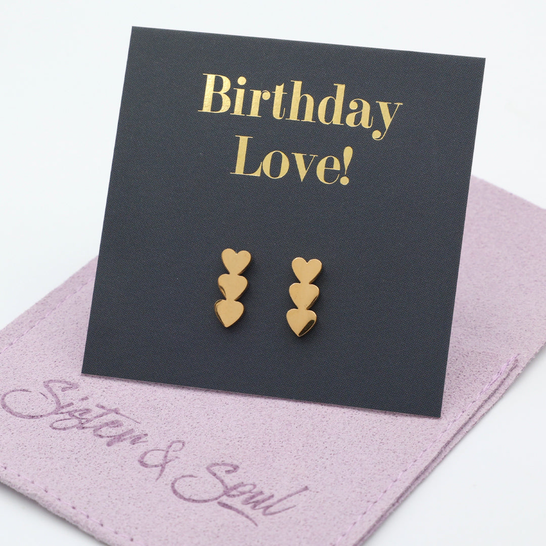 A pair of gold heart stud earrings displayed on a black card that reads 'Birthday Love!' in gold lettering, resting on a purple fabric pouch with the words 'Sister & Soul' in script.