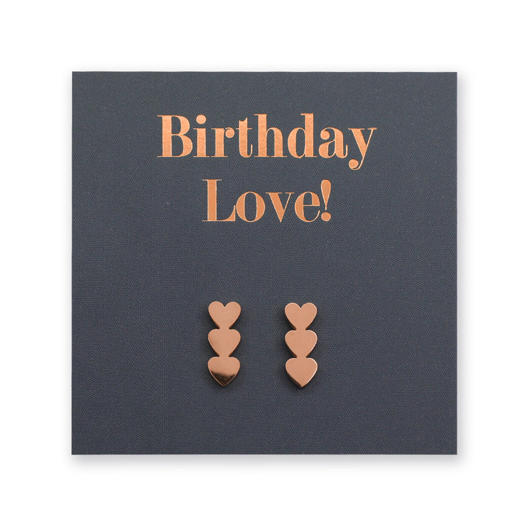 A pair of rose gold heart-shaped stud earrings displayed on a dark gray square gift card with the words 'Birthday Love!' in rose gold lettering.