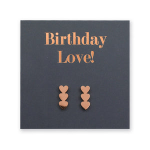 A pair of rose gold heart-shaped stud earrings displayed on a dark gray square gift card with the words 'Birthday Love!' in rose gold lettering.