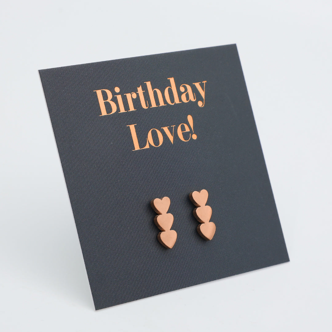 A pair of rose gold heart stud earrings displayed on a dark charcoal gray card that reads 'Birthday Love!' in rose gold lettering, set against a clean white background.