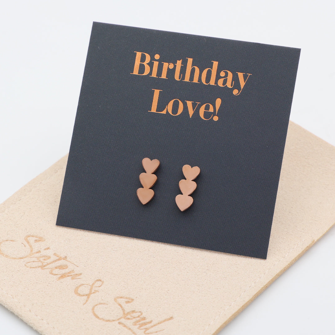 Three-pair heart stud earrings in a rose gold tone, displayed on a black square card that reads 'Birthday Love!' in rose-gold lettering, resting on a beige pouch with the script 'Sister & Soul'.
