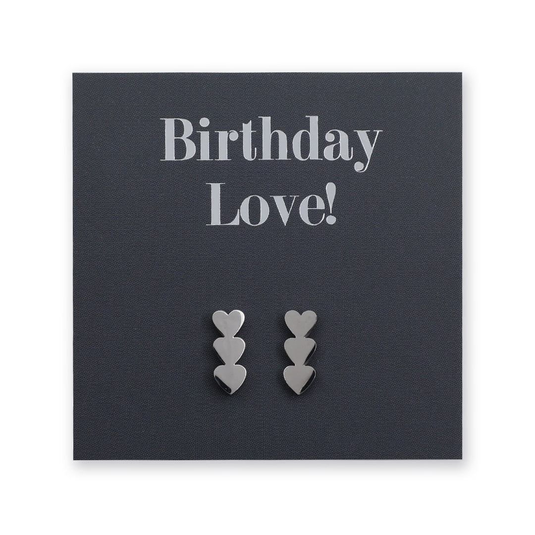A pair of silver heart-shaped stud earrings displayed on a dark charcoal card that reads 'Birthday Love!'.