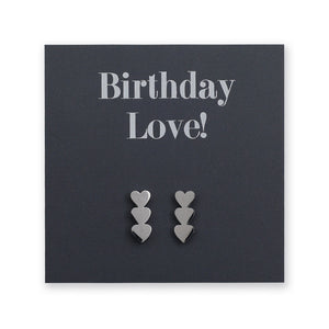 A pair of silver heart-shaped stud earrings displayed on a dark charcoal card that reads 'Birthday Love!'.